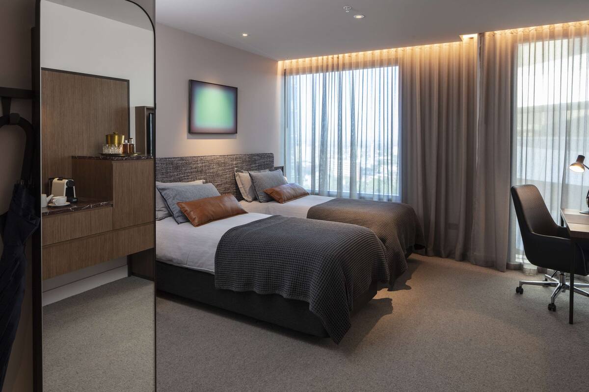 A hotel room with two beds, a desk, a chair, and a large window with curtains.