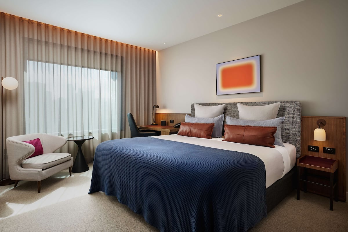 A modern hotel room with a large bed, a chair, a small table, and a wall-mounted lamp.