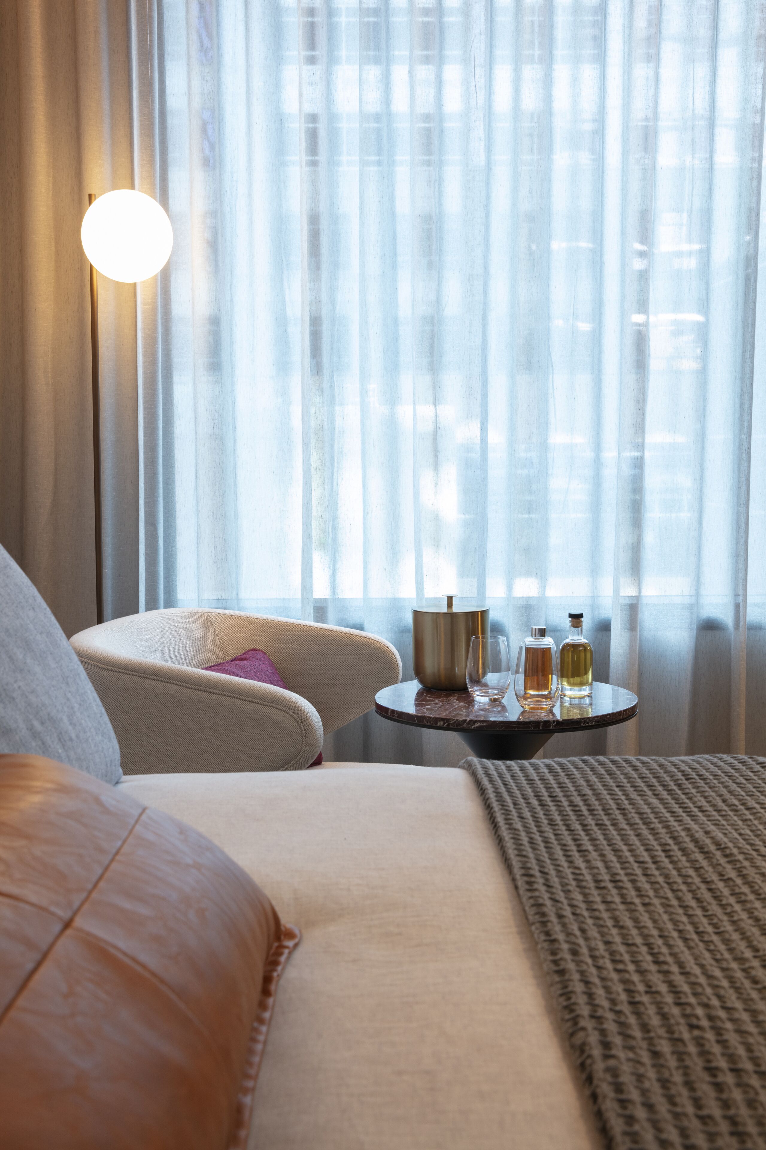 Habitación de hotel con cama, una silla, una mesa con lámpara y ventana con cortinas translúcidas.