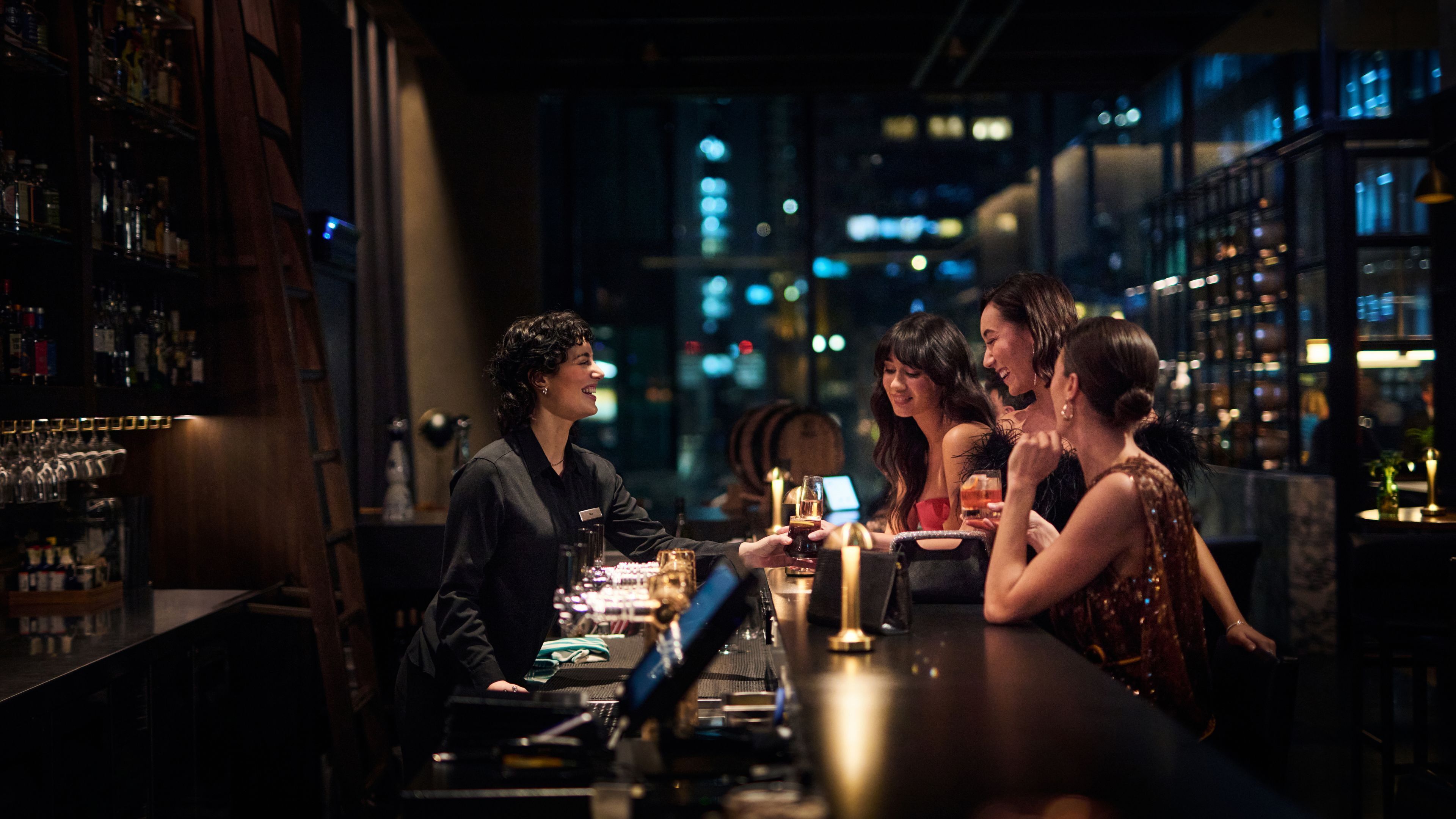 Een bar met een barman en gasten. Het interieur heeft een modern design met een donker kleurenschema. Er zijn planken met flessen en glazen. Op de achtergrond is een stadsbeeld bij nacht te zien.