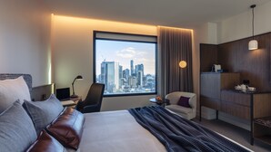 A modern hotel room with a large bed, a desk, a chair, a small table, and a view of the city skyline. - Next Hotel Melbourne, Curio Collection by Hilton (Melbourne)