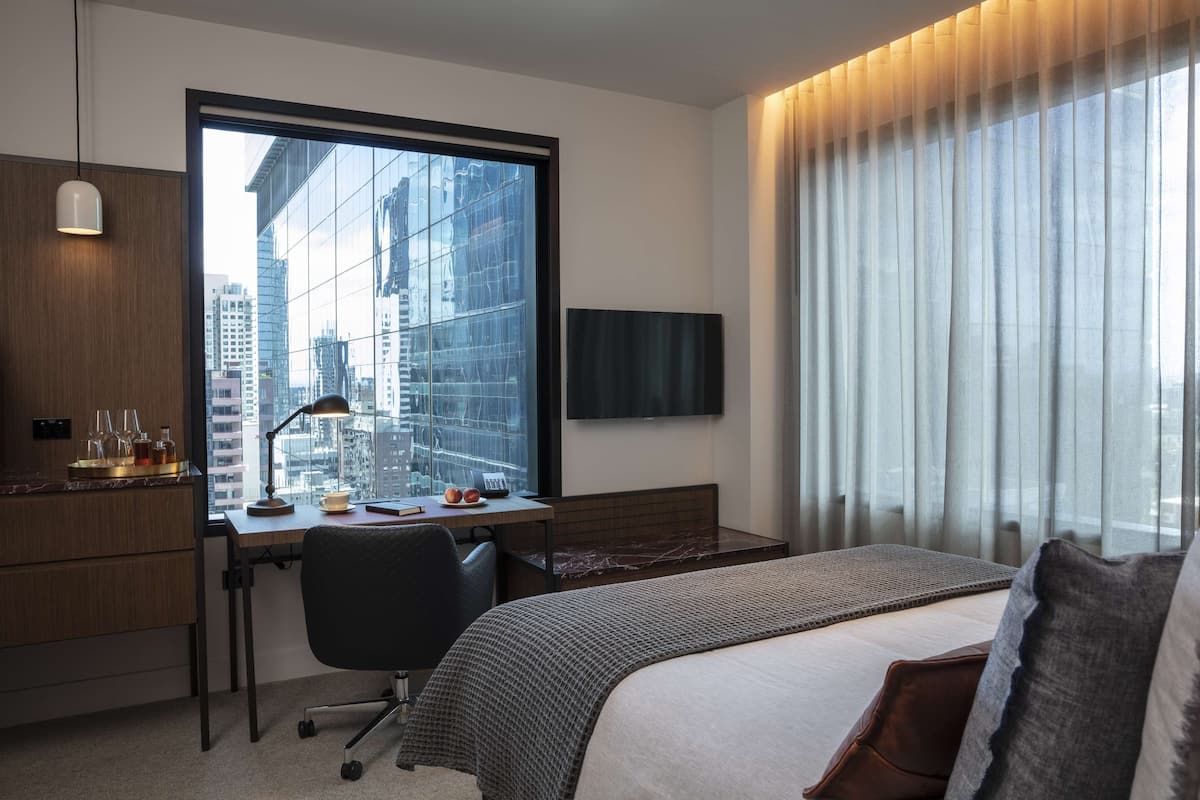 A modern hotel room with a large window, a desk with a computer, a bed, and a TV mounted on the wall.
