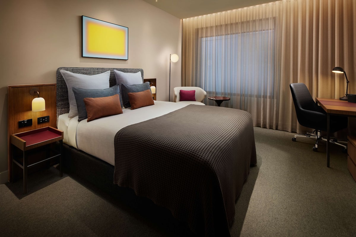 A hotel room with a large bed, a desk, a chair, a lamp, and a window with curtains.