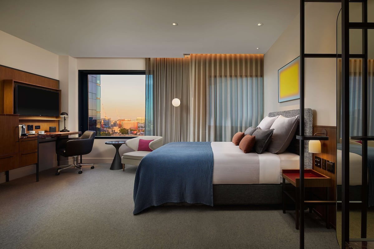 A modern hotel room with a bed, desk, chair, and a large window offering a city view.
