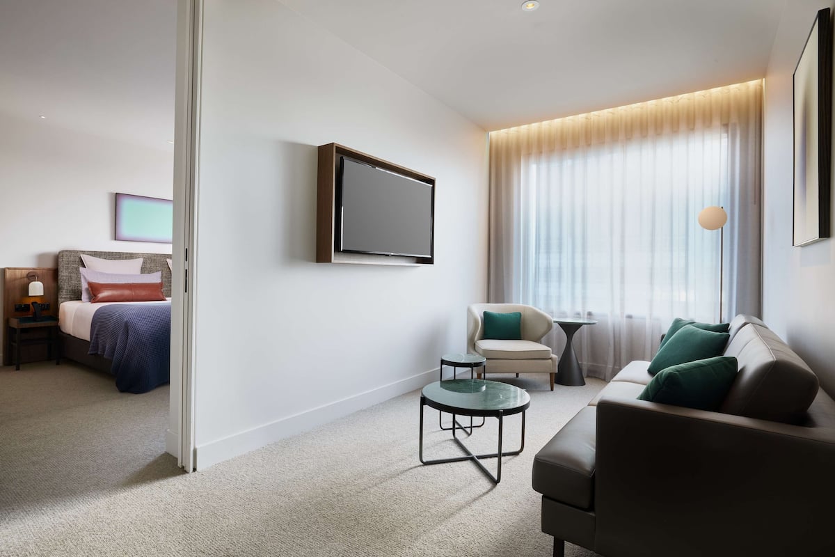 A modern hotel room with a bed, a sofa, a small table, and a TV mounted on the wall.