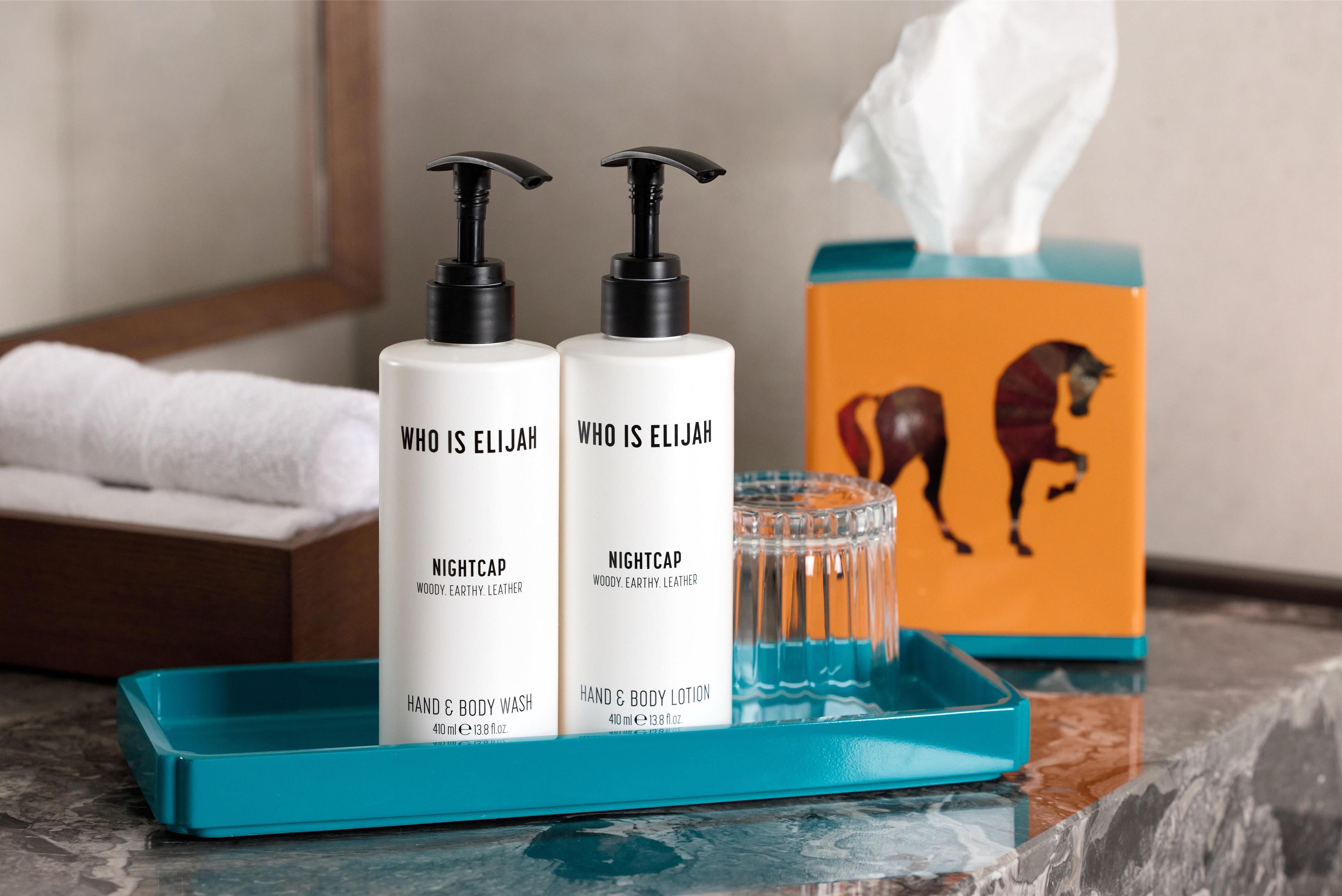 Two bottles of Who is Elijah Nightcap body wash and lotion, a tissue box, and a glass of water on a blue tray.