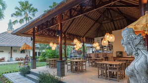 Un espacio de comedor al aire libre con mesas y sillas de madera, techo de paja y lámparas de mimbre colgantes.