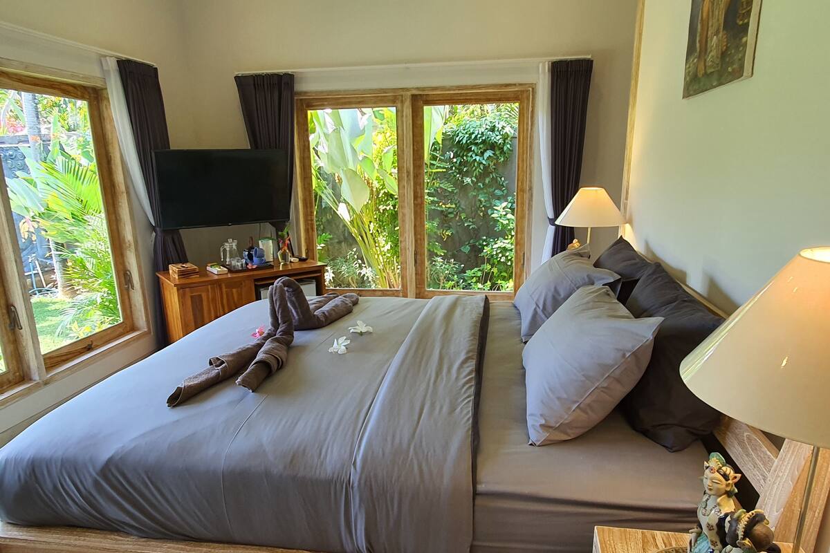 Un dormitorio con una cama grande, un televisor, una mesita de noche y vistas a la vegetación a través de las ventanas.
