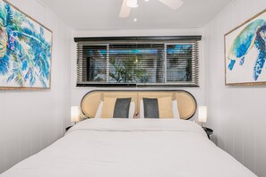 Una cama bien hecha con una cabecera de ratán, dos lámparas de noche y una ventana con persianas.