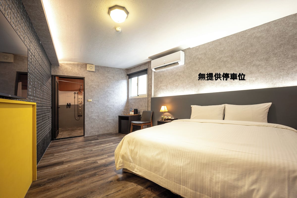 Quarto de hotel moderno com uma cama grande, uma escrivaninha e um banheiro visível através de uma porta aberta.