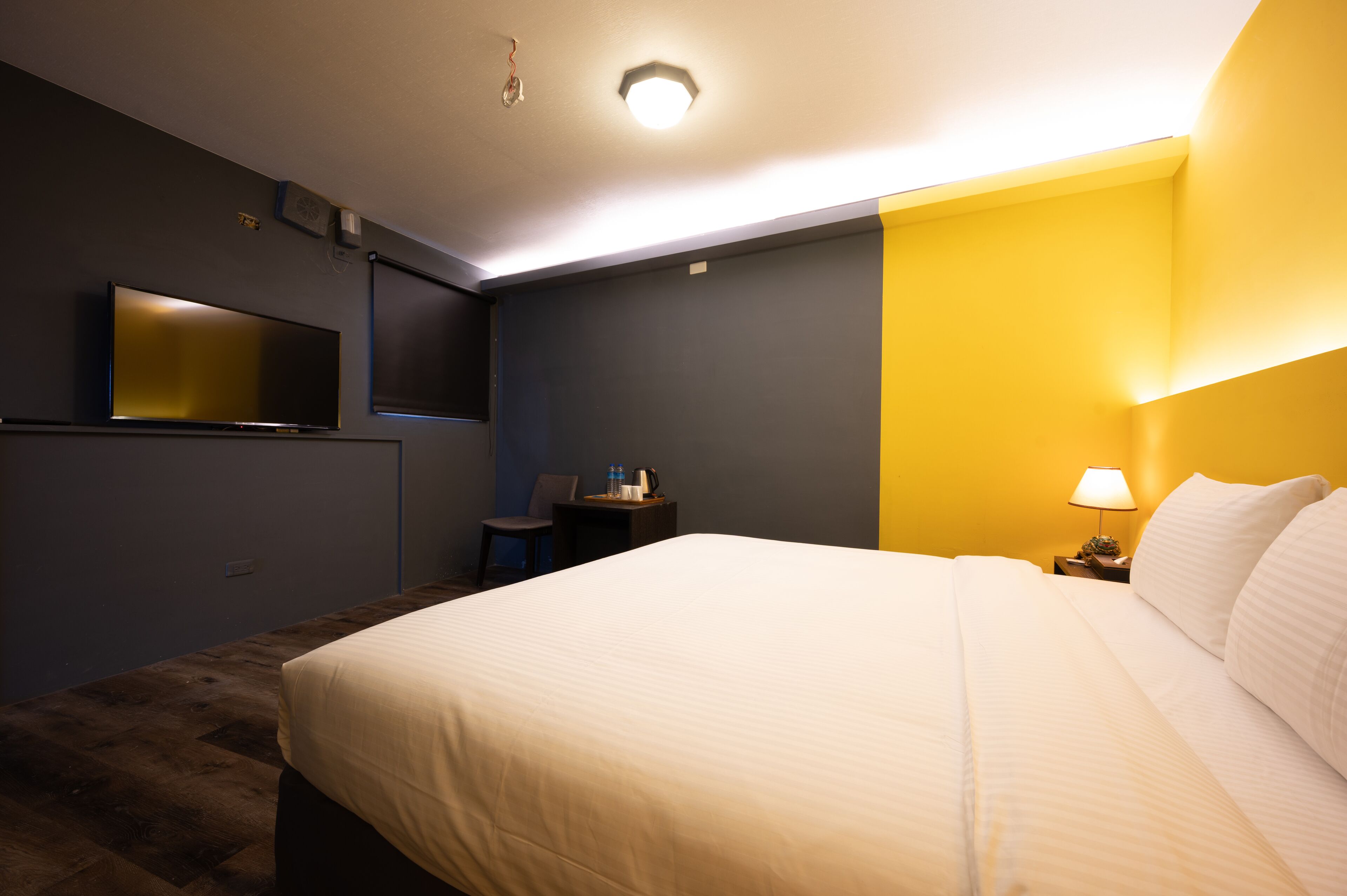 A hotel room with a large bed, a desk with a chair, a flat-screen TV mounted on the wall, and a lamp on the nightstand.