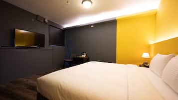 A hotel room with a large bed, a desk with a chair, a flat-screen TV mounted on the wall, and a lamp on the nightstand.