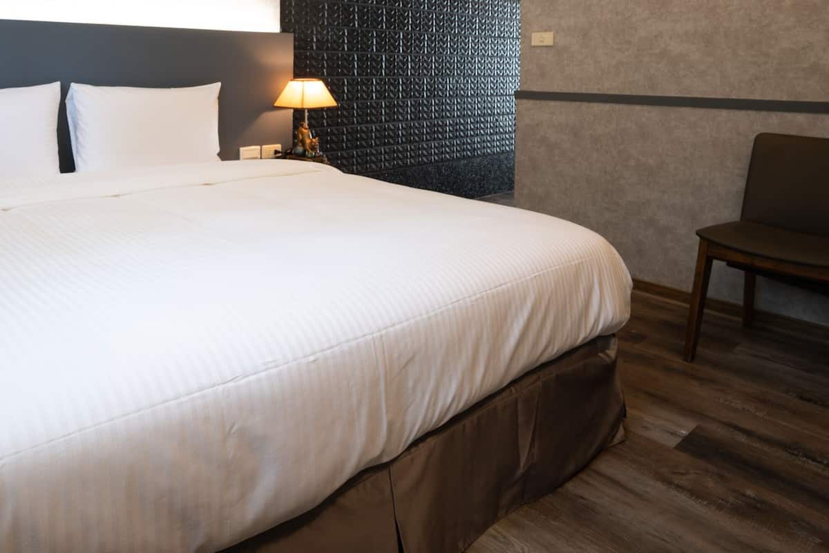Quarto de hotel com uma cama grande, uma mesa com luminária, uma cadeira e um dispensador de água.