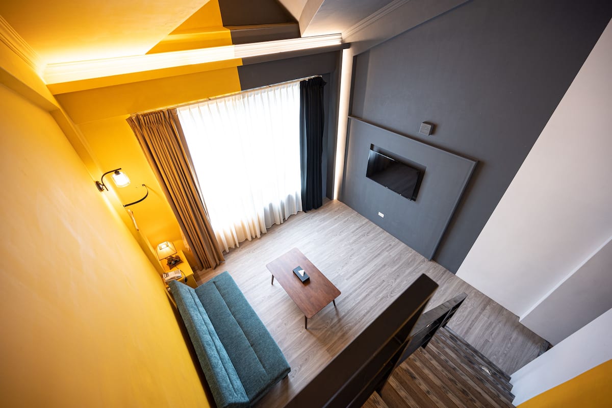 Quarto de hotel moderno com piso de madeira, sofá, mesa de centro, televisão fixada na parede e janela com cortinas.