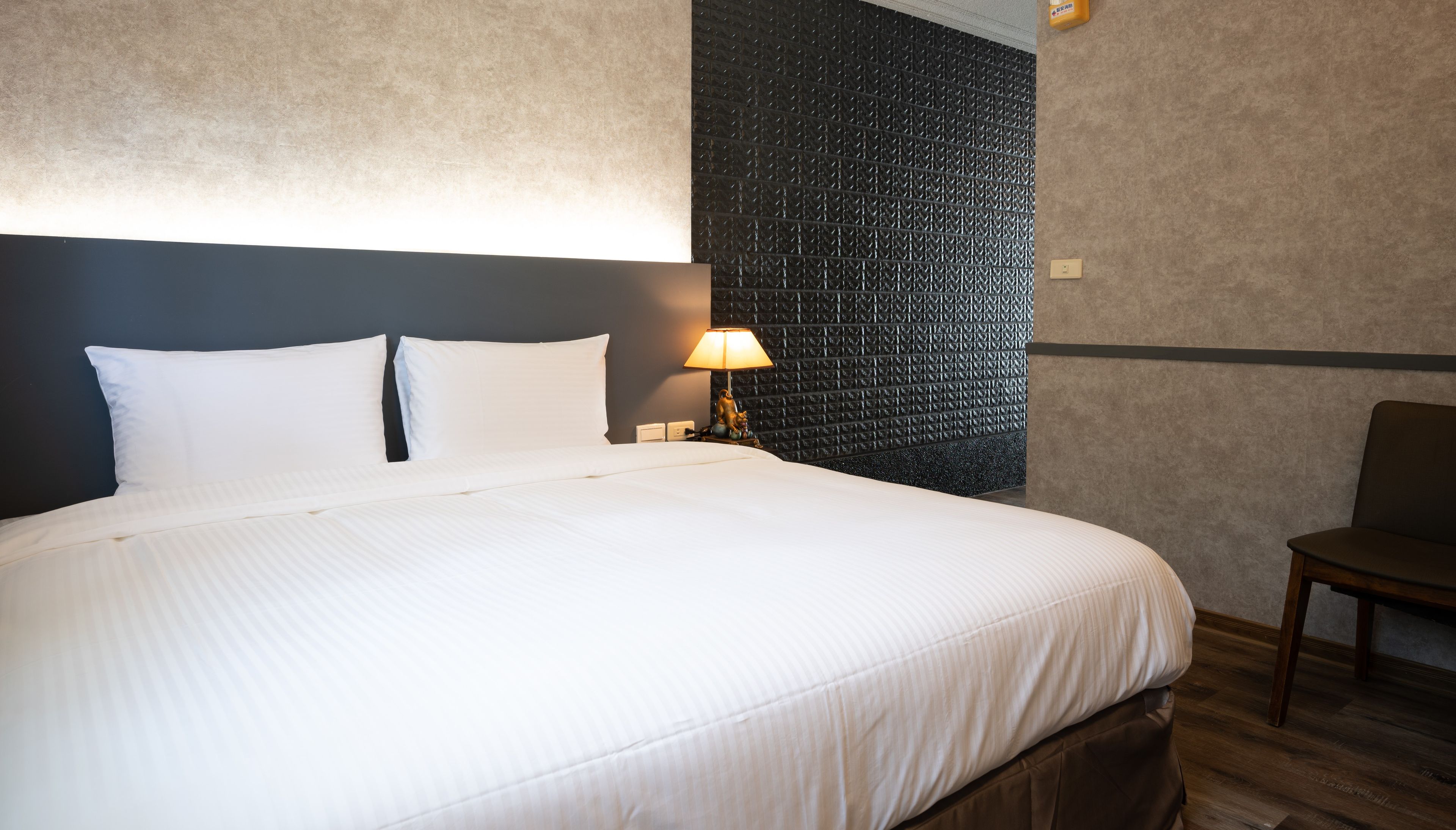 A hotel room with a large bed, a nightstand with a lamp, and a chair. The room has a textured wall and a ceiling fan.