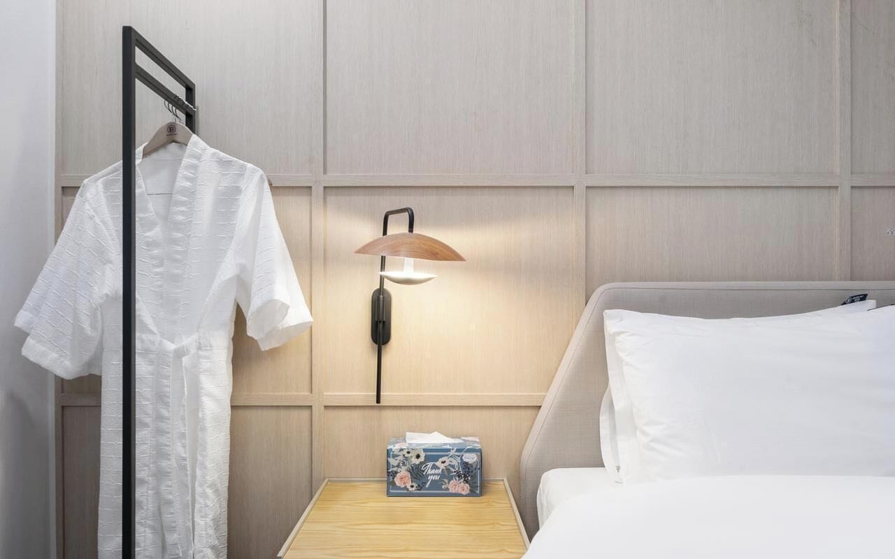 Ein Hotelzimmer mit einem Bett, einem Nachttisch mit Lampe, einem aufgehängten Bademantel und einer kleinen Schachtel mit Blumenmuster.