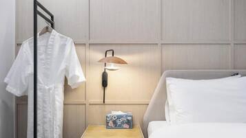 A hotel room with a bed, a bedside table with a lamp, a hanging robe, and a small box with a floral design.