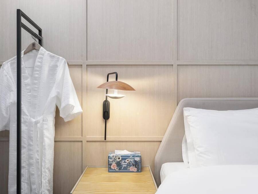 A hotel room with a bed, a bedside table with a lamp, a hanging robe, and a small box with a floral design.