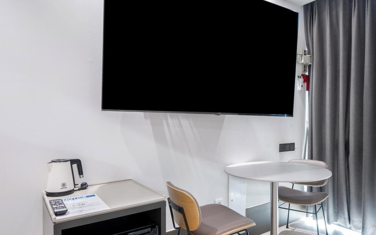 Habitación de hotel con televisión de pantalla plana, una mesita con hervidor, una silla y un escritorio con control remoto.