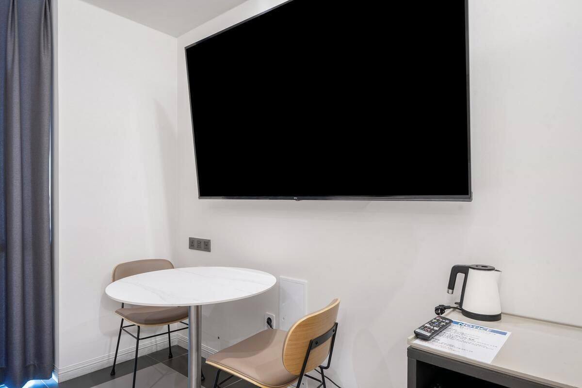 Una habitación con una mesa redonda, dos sillas, una pantalla de proyector, un hervidor y una pequeña mesa con un papel.