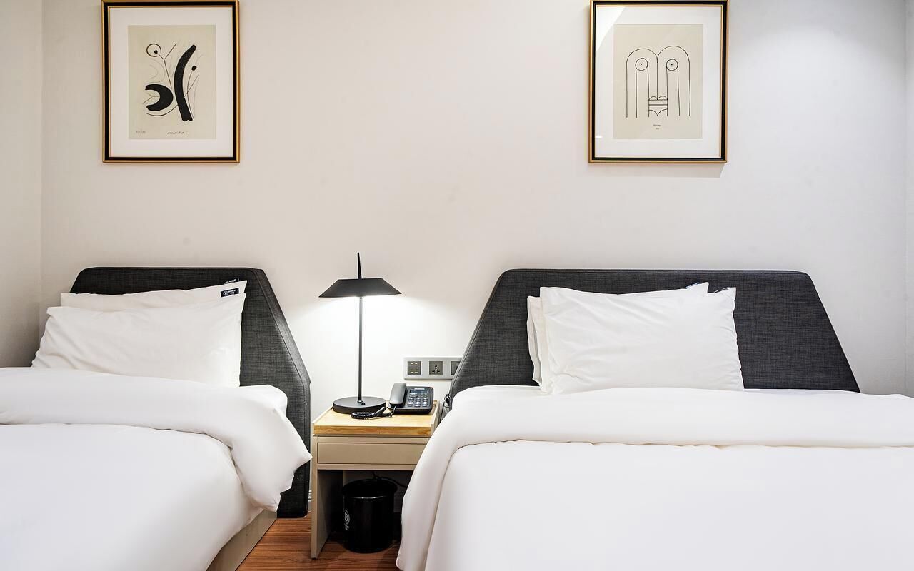A hotel room with two beds, a bedside table with a lamp, a phone, and a trash bin.