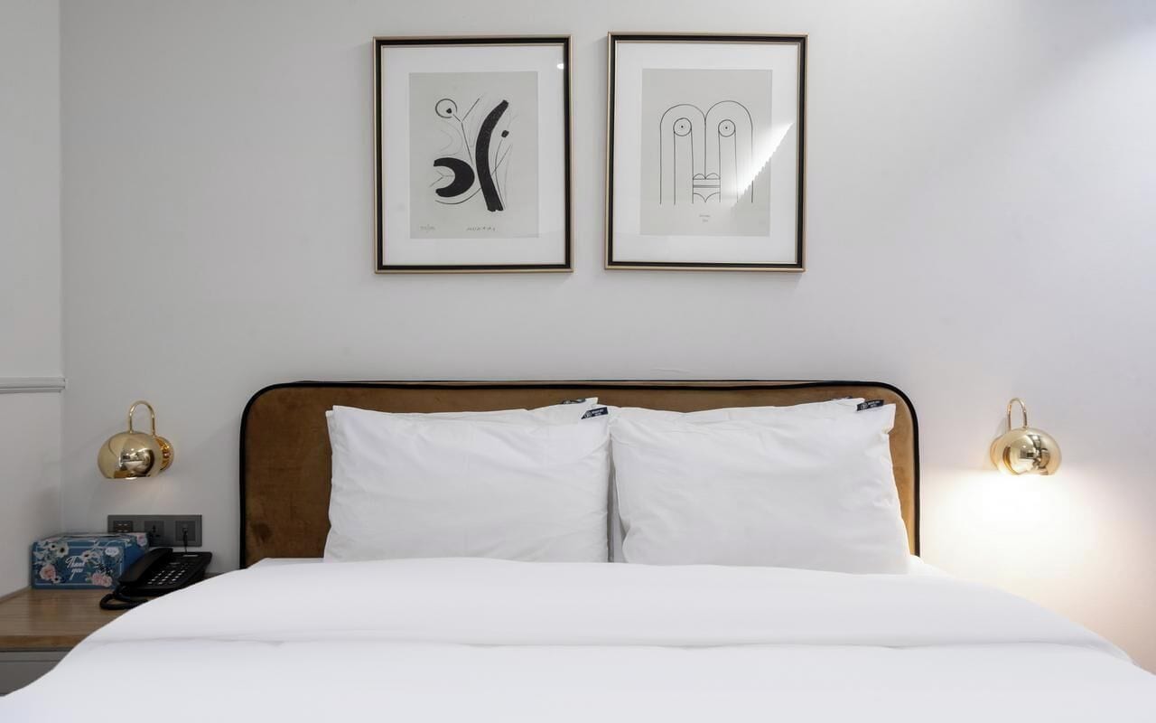 Una camera d'albergo con testiera in legno, biancheria da letto bianca, due opere d'arte incorniciate appese al muro, lampade da comodino e un piccolo tavolino con un telefono e una scatola decorativa.