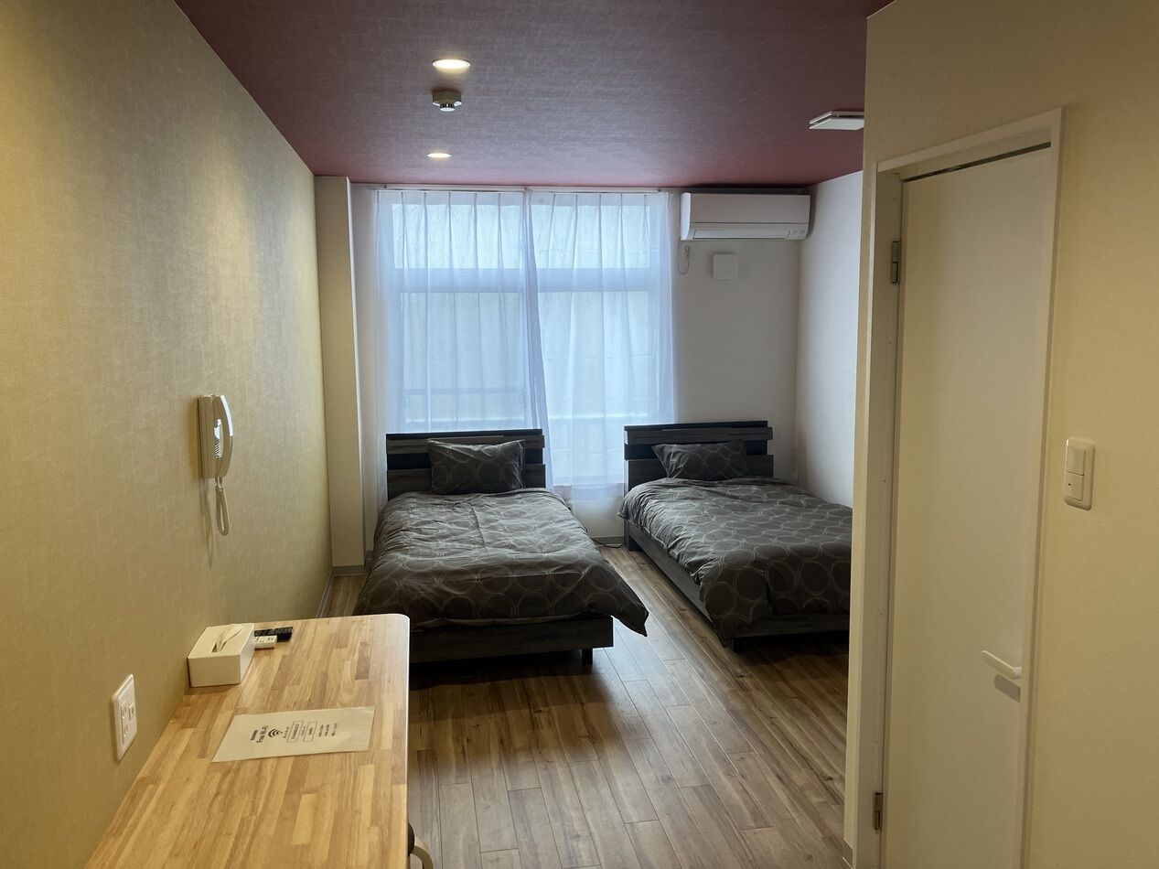 A small, clean room with two beds, a wooden desk, a phone, and a window with sheer curtains.