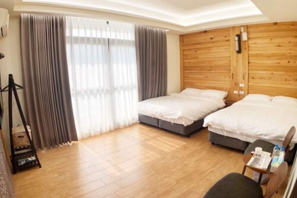A hotel room with a wooden ceiling, two beds, a small table with a chair, and a window with curtains.