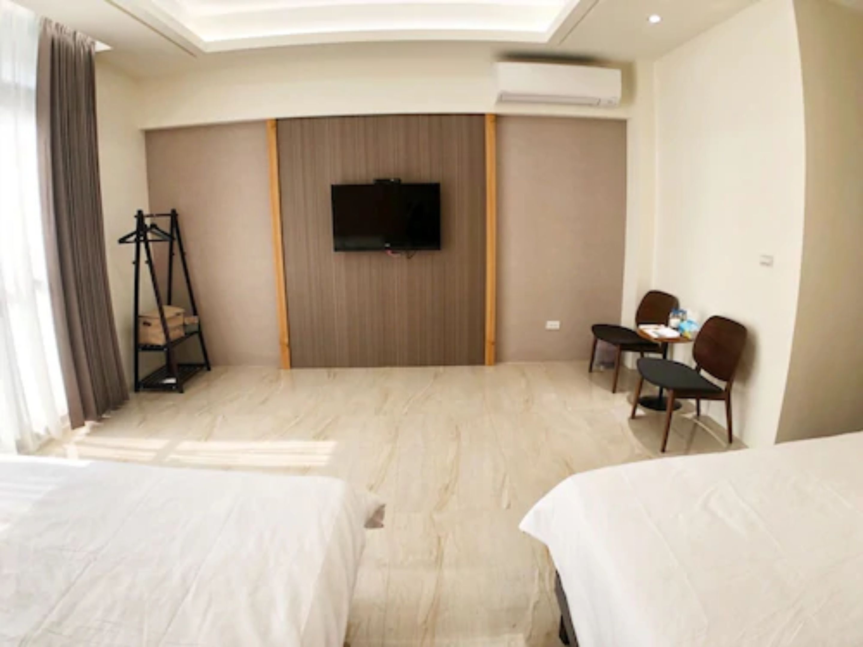 A hotel room with two beds, a TV mounted on the wall, a small table with chairs, and an air conditioning unit.