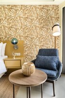 A hotel room with a patterned wallpaper, a bed with white bedding, a nightstand with a lamp, a blue armchair, and a small wooden table with a decorative item.