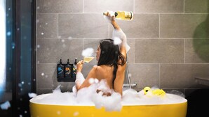 Una persona en una bañera, rodeada de burbujas, con botellas de champán y una copa.