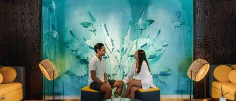 Una pareja sentada en un sofá en una habitación con un gran mural de peces y corales. Hay dos lámparas de pie y una pequeña mesa con una planta.
