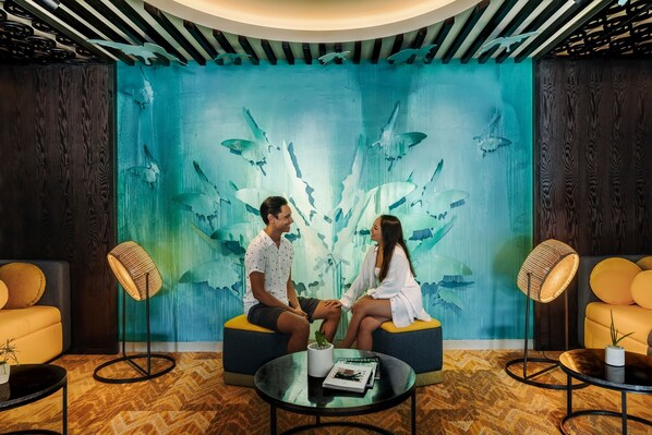 Una pareja sentada en un sofá en una habitación con un gran mural de peces y corales. Hay dos lámparas de pie y una pequeña mesa con una planta.