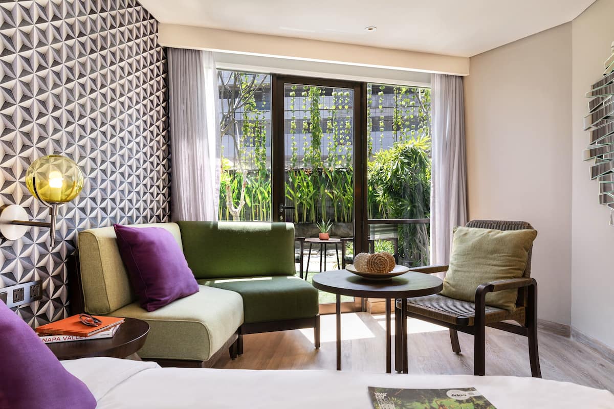 Een moderne hotelkamer met een groene bank, een houten stoel, een klein tafeltje en een groot raam met uitzicht op groen.