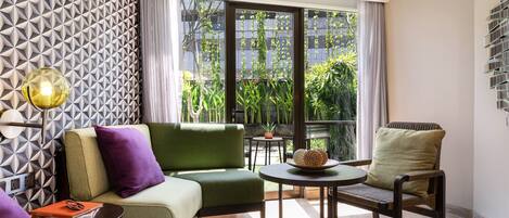 A modern hotel room with a green sofa, a wooden chair, a small table, and a large window offering a view of greenery outside.