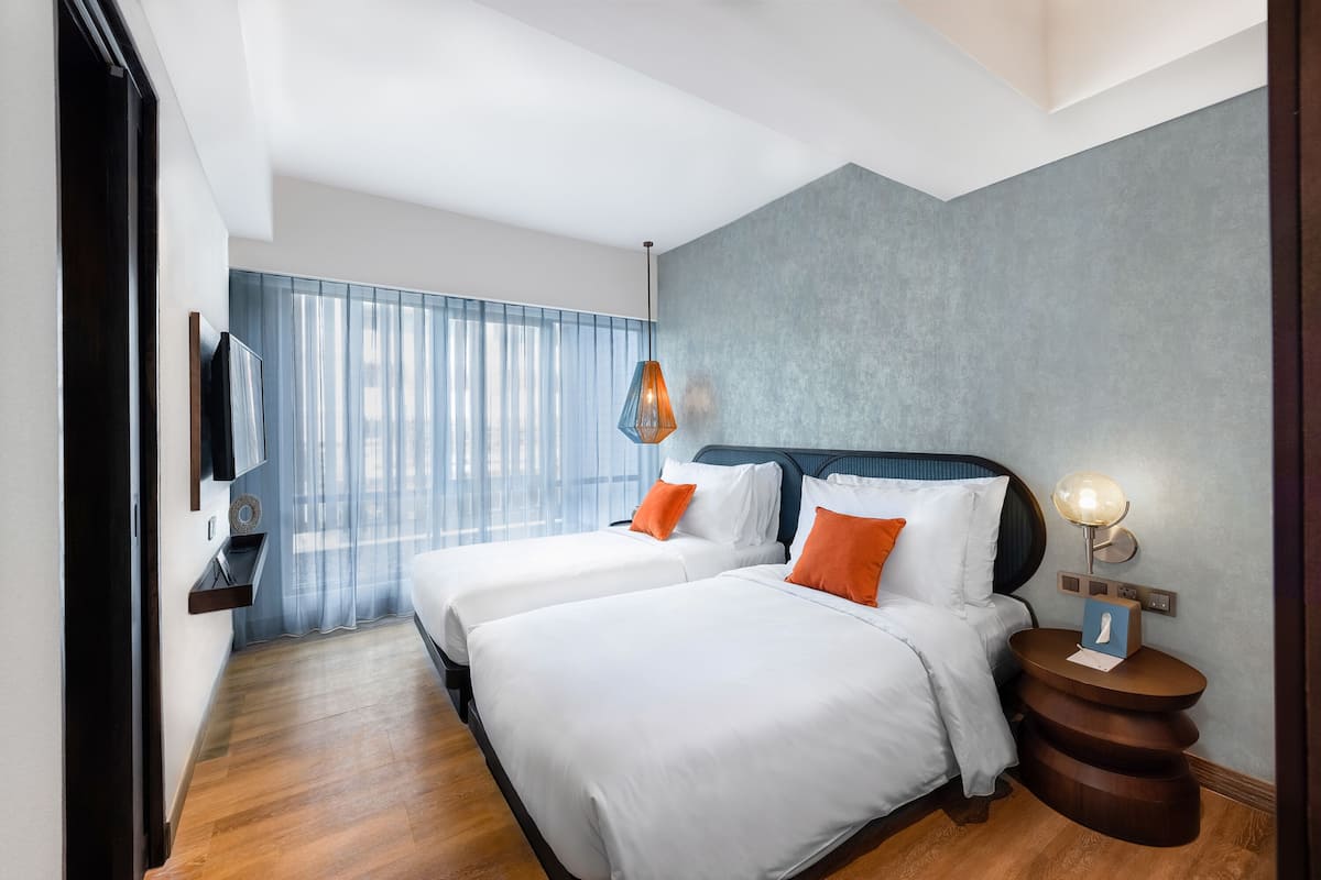 Een hotelkamer met twee bedden, een televisie, een nachtkastje met lamp en een raam met dunne gordijnen.