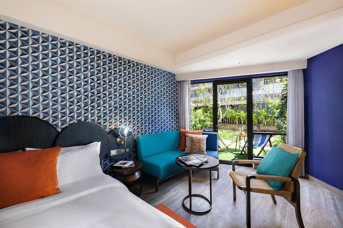 Een hotelkamer met een bed, een bank, een stoel, een klein tafeltje en een balkon met planten.