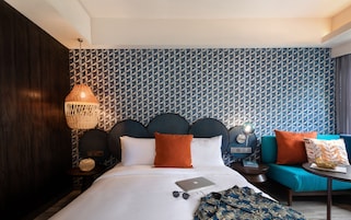 A modern hotel room with a geometric patterned wallpaper, a large bed with white bedding, a laptop, sunglasses, and a teal armchair.