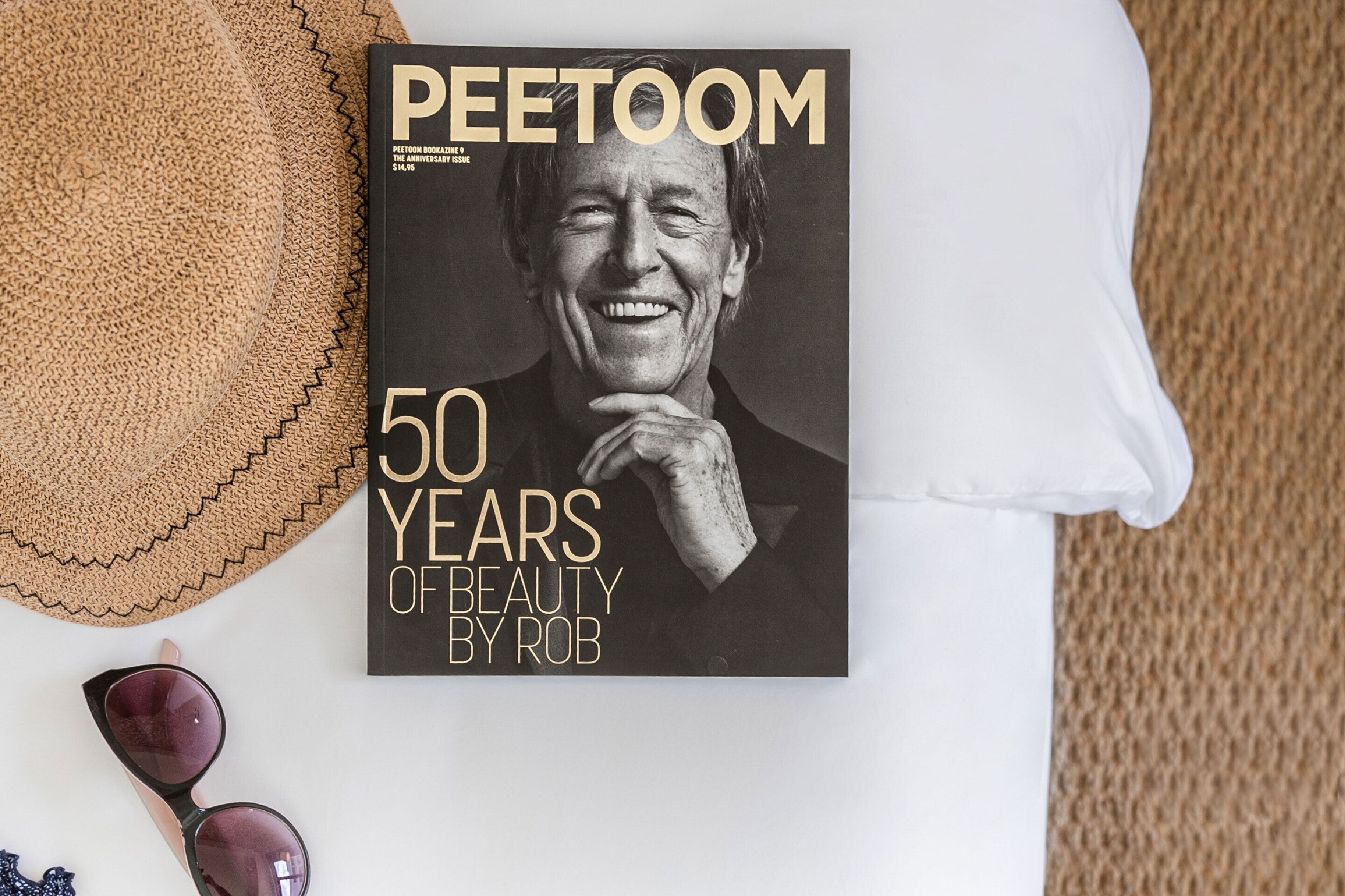 Un livre intitulé « PEETOOM, 50 ANS DE CRÉATIONS DE ROB », posé sur une surface blanche, avec un chapeau tressé et des lunettes de soleil à côté.