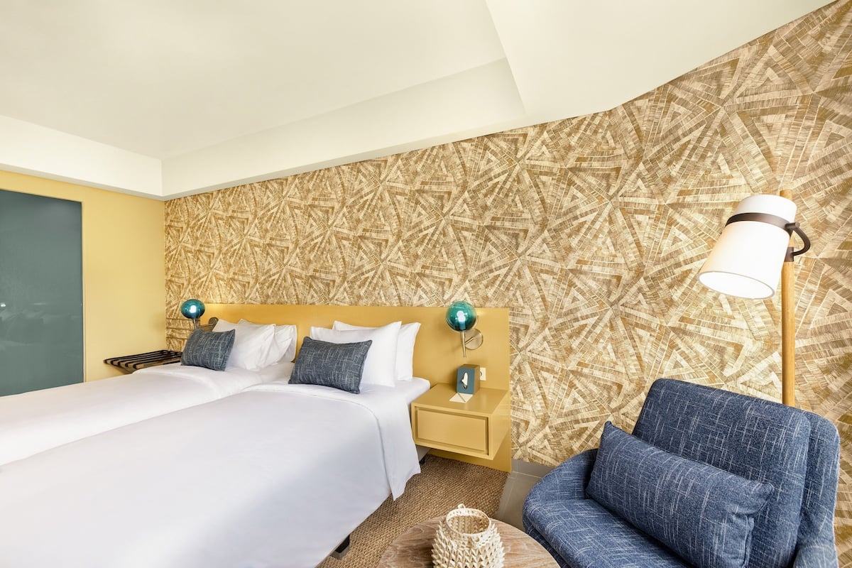 Hotelkamer met een groot bed, nachtkastje met lamp, een blauwe fauteuil en een behang met patroon.