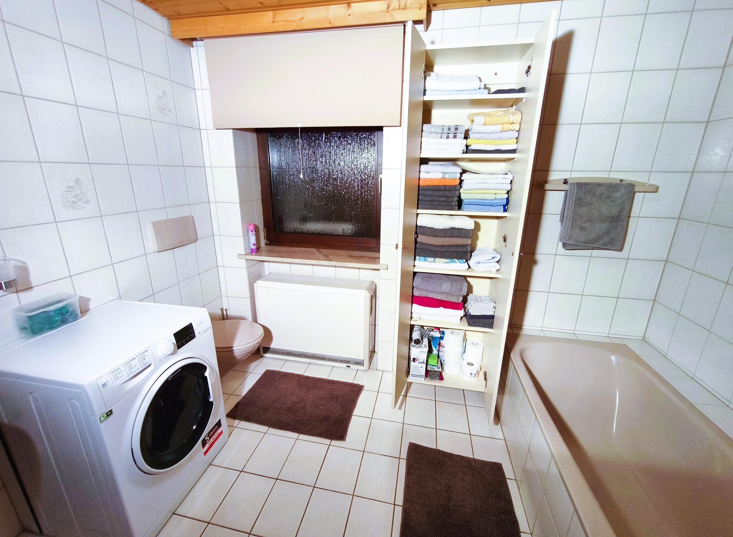 A compact laundry room with a washing machine, a radiator, a window, and a towel rack.