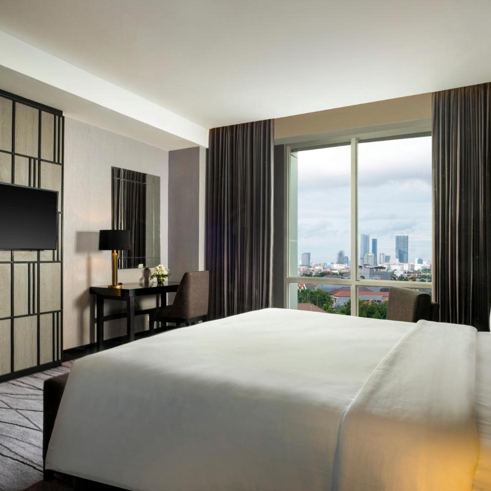 A hotel room with a large bed, a desk with a lamp, a chair, and a view of a cityscape through a large window.