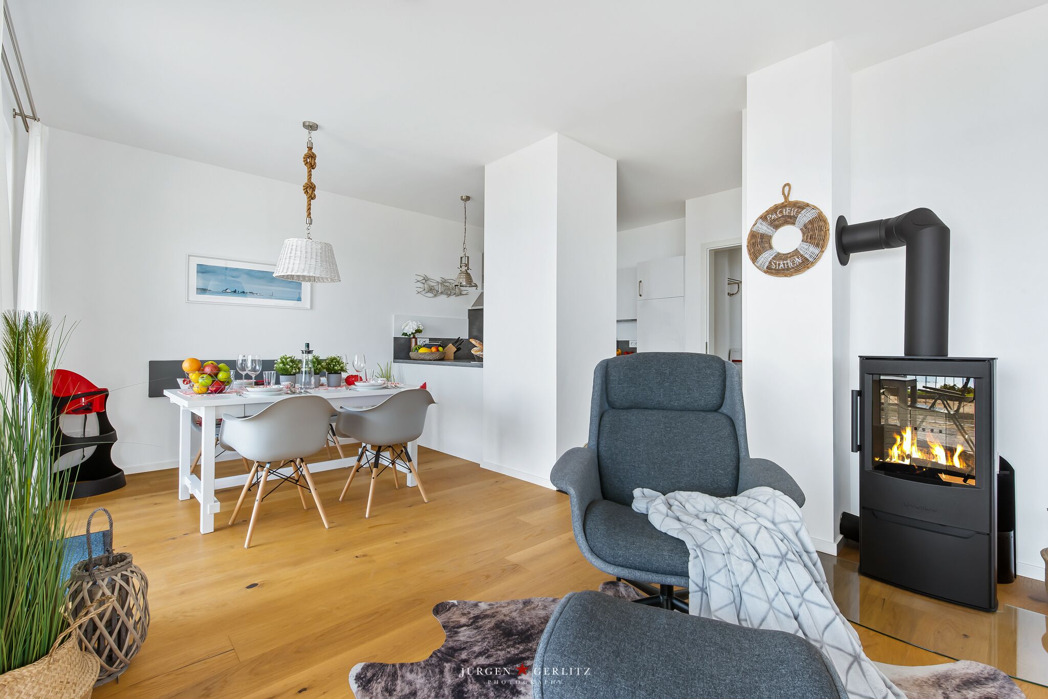 A modern living room with a grey armchair, a dining area with a white table and chairs, and a black wood-burning stove.