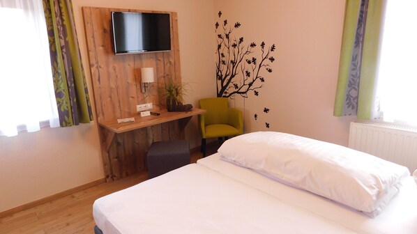Una habitación de hotel con una cama, un escritorio con una silla, un televisor montado en la pared y un adhesivo decorativo de árbol.