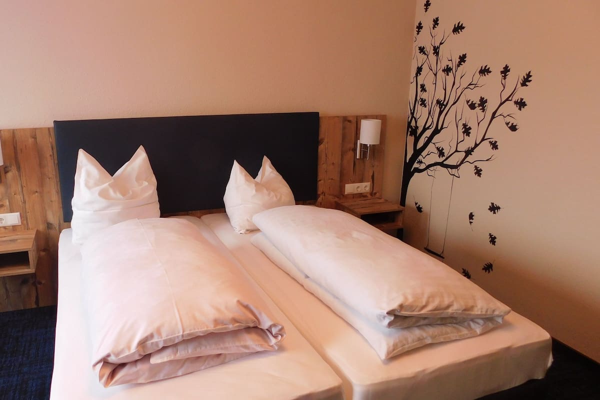 Ein Doppelbett mit zwei Kissen, ein Holz-Kopfbrett, Nachttischlampen und ein Wandsticker mit Baummotiv.