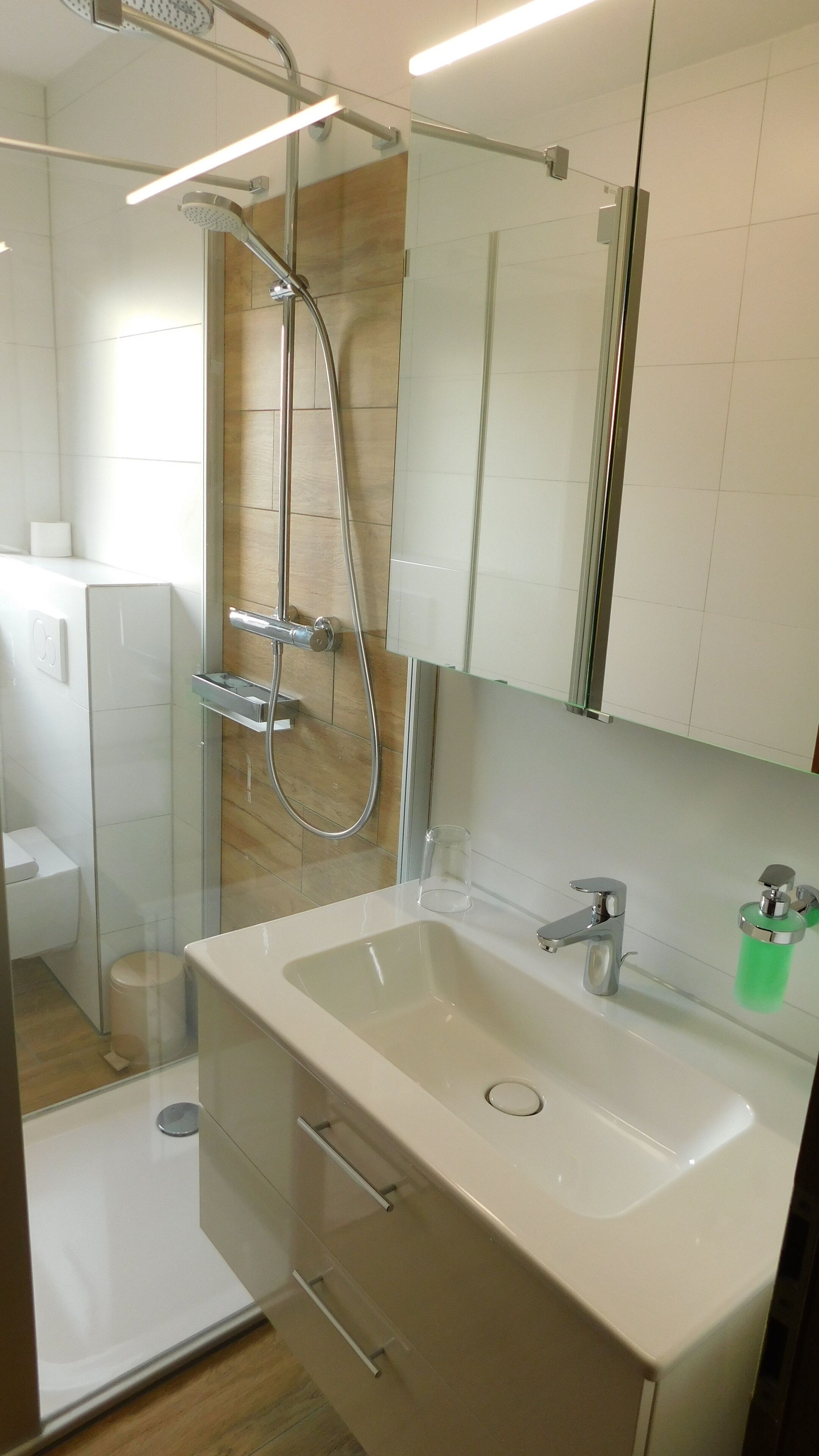 Ein modernes Badezimmer mit einer Duschkabine aus Glas, einem weißen Waschbecken mit grünem Seifenspender und einem Spiegel darüber.