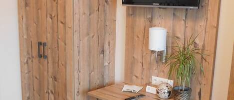 Una habitación con un armario de madera, un televisor de pantalla plana montado en un panel de madera, una mesita con una lámpara, una planta en maceta y un otoman gris.