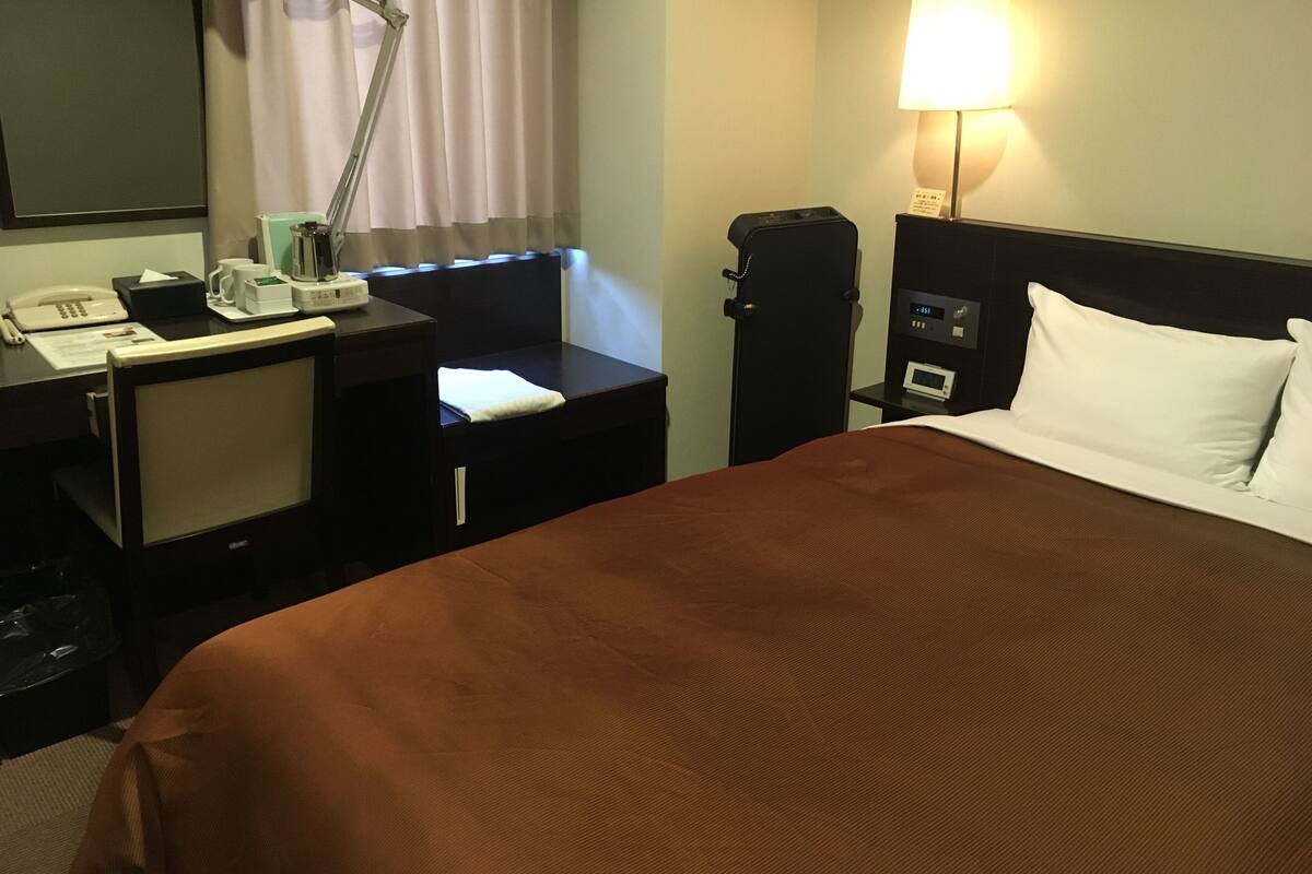 A hotel room with a bed, a desk with a lamp, a telephone, a coffee maker, and a nightstand with a clock.