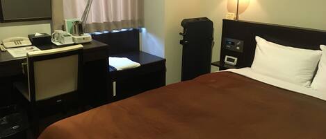 A hotel room with a bed, a desk with a lamp, a telephone, a coffee maker, and a nightstand with a clock.