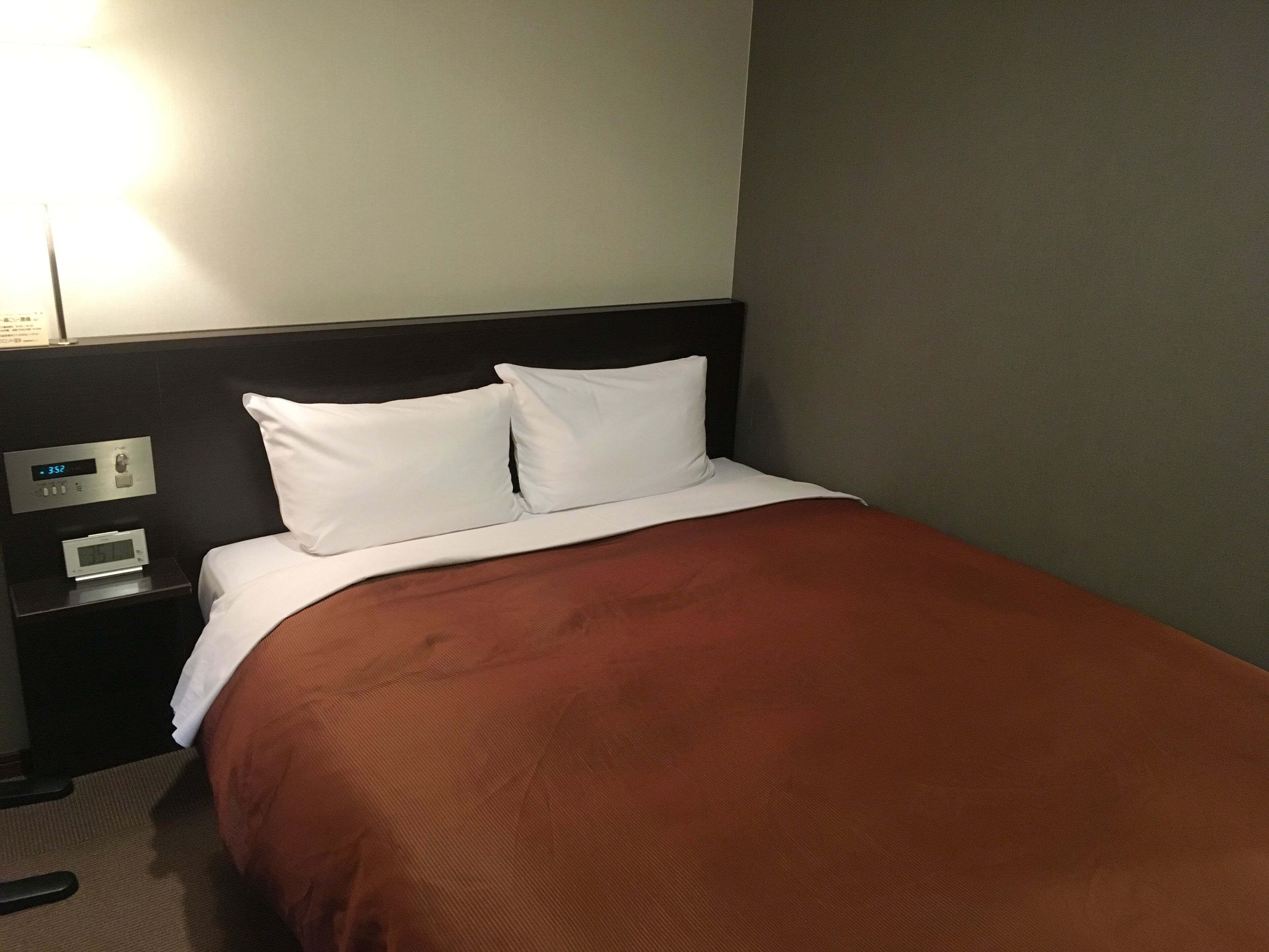 Una habitación de hotel con cama, almohadas blancas, colcha marrón, mesita de noche con reloj digital y una lámpara de pared.